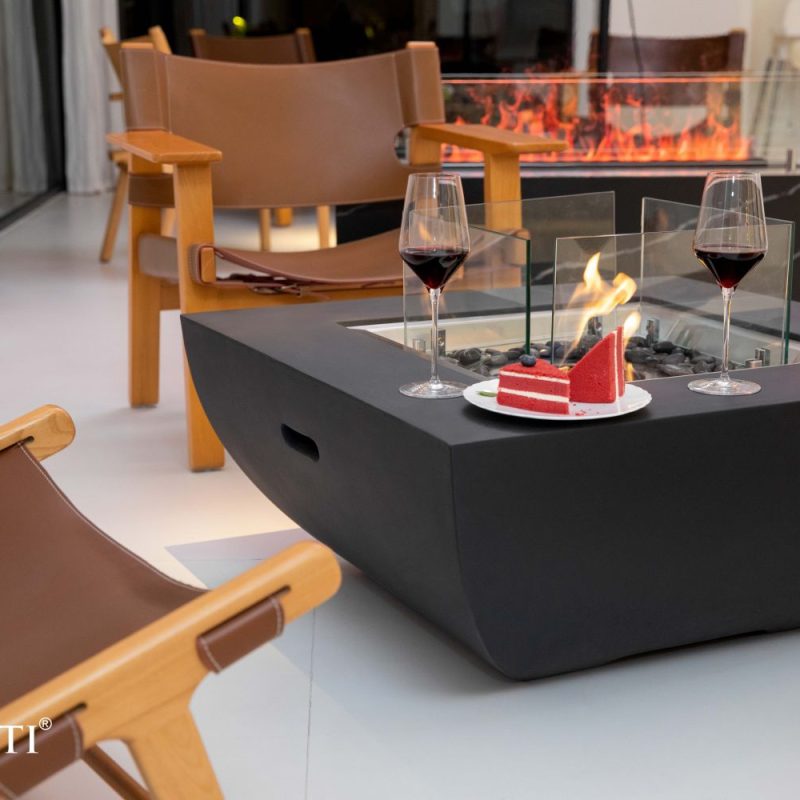 Elementi Aurora ECG08 black ethanol fire pit coffee table with glass wind guard and active flame, styled with slice of red velvet cake, two wine glasses, and brown leather lounge chairs for outdoor entertaining
